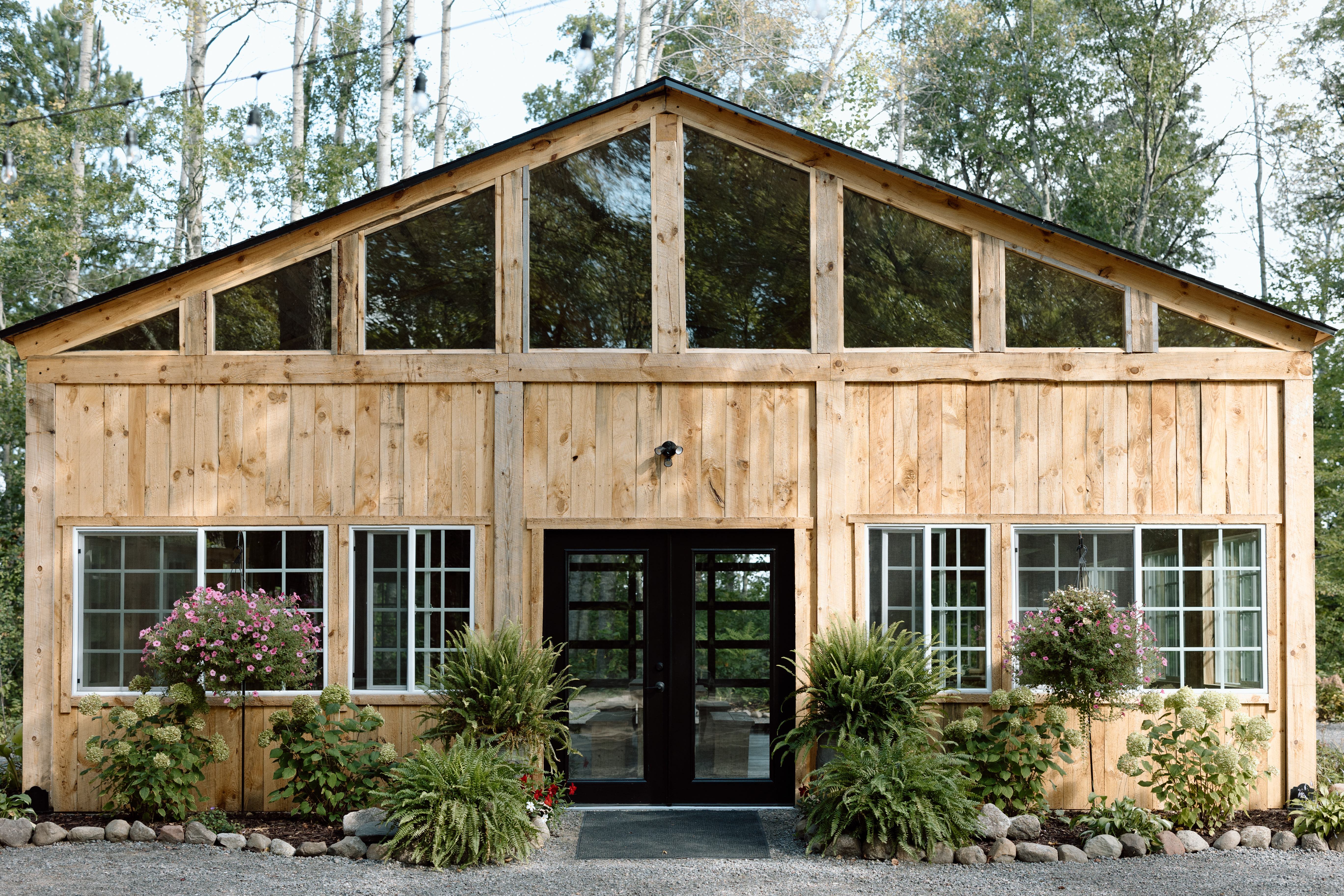 The Emerald Cottage, a timber-framed venue with a glass gable peak surrounded by tall trees, string lights, and flowering gardens
