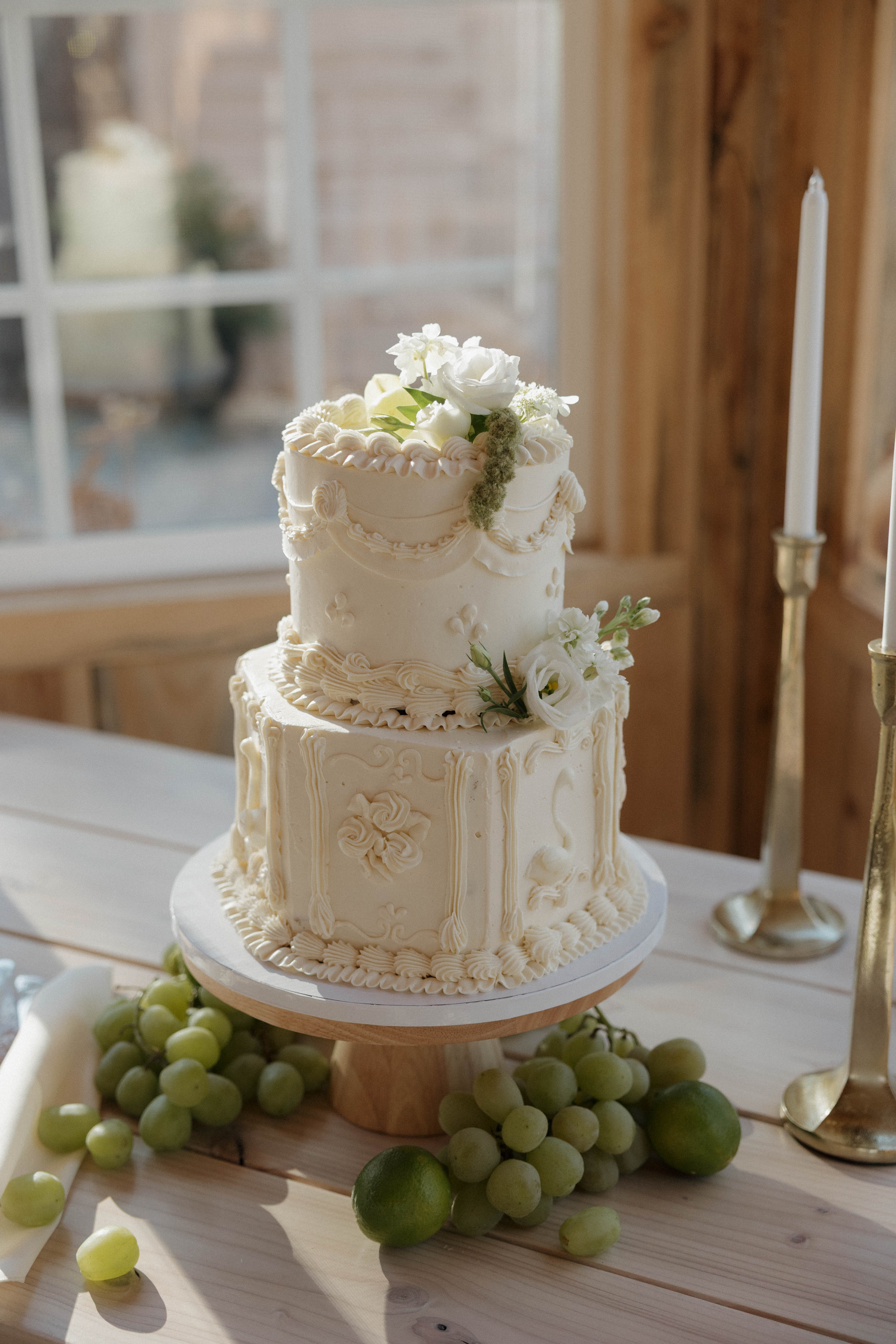 A vintage-style two-tier buttercream wedding cake with white roses, styled with grapes and brass candlesticks in the cottage barn