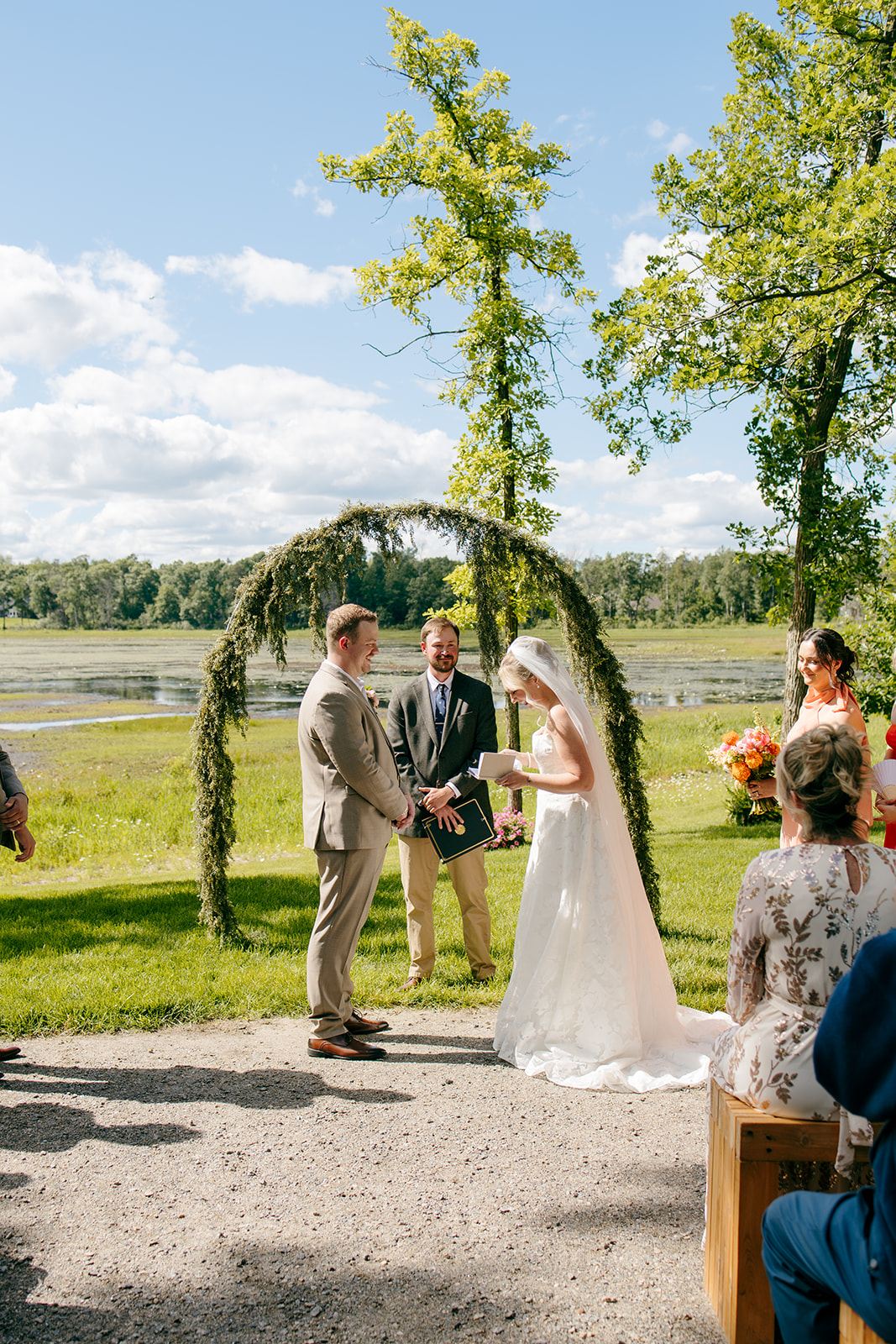 A couple exchanging vows beneath the greenery-covered lakeside arch with the officiant, guests, and lake behind them