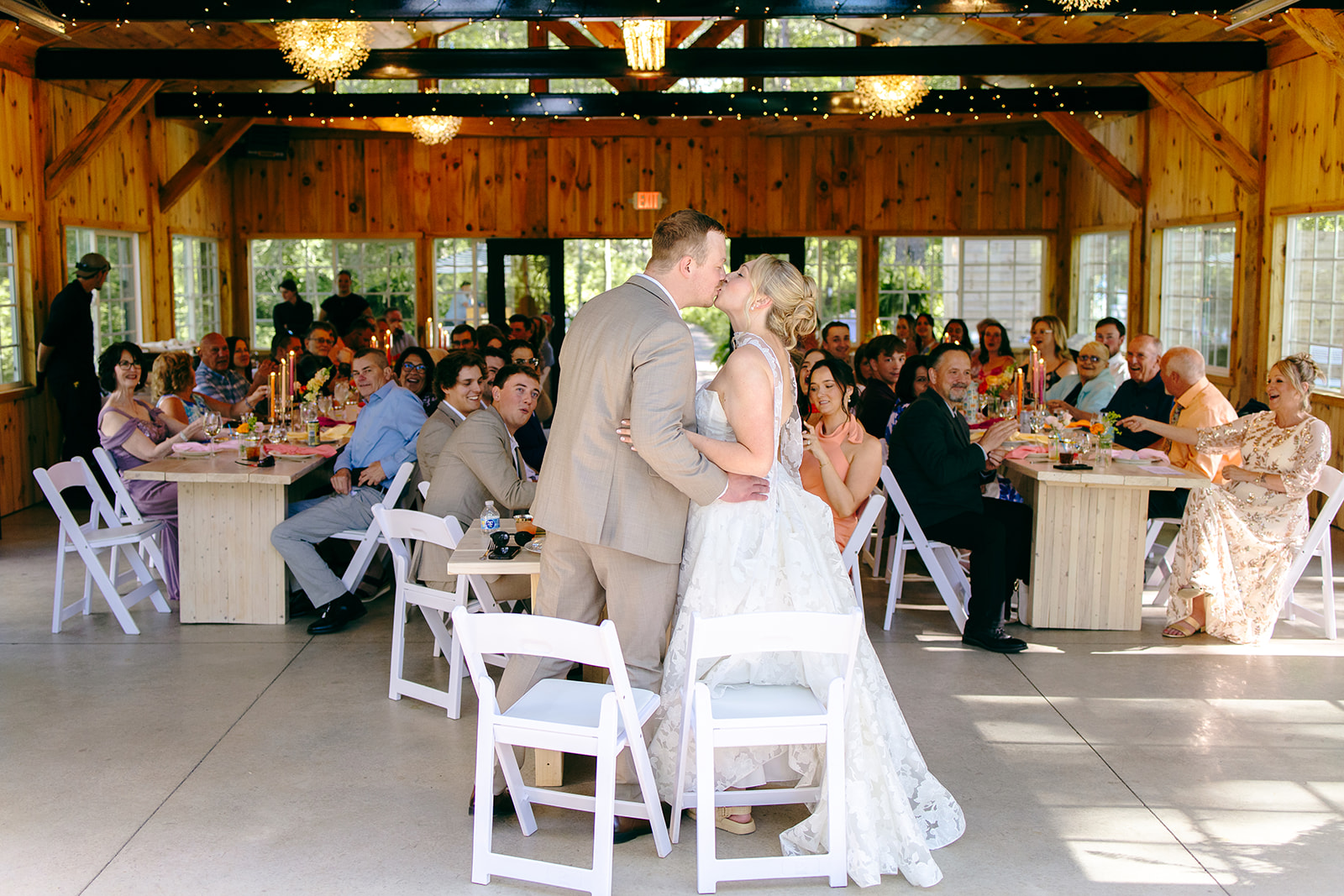 A couple sharing a kiss surrounded by seated guests at candlelit farm tables inside the timber cottage